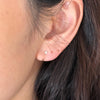 A close-up of a person's ear wearing a small gold huggie hoop earring and a tiny gold stud earring with a clear gemstone.