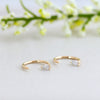 A pair of gold open hoop earrings with small marquise-cut clear stones, displayed on a light surface with soft-focus white flowers in the background.
