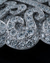 A close-up of an ornate silver-toned jewelry piece, likely a brooch or decorative pendant, heavily set with clear round and baguette diamonds against a solid black background.