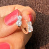 A close-up of a hand with pink nail polish holding a pair of rose gold huggie hoop rings with triple-cut clear gemstones, set against a dark brown textured fabric background.
