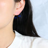 Close-up of a person's ear wearing two small stud earrings, with blue text labels 'LG' and 'SM' near the studs. The person has dark hair and is wearing a white top against a light background.