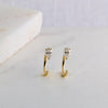 A pair of gold huggie hoop earrings with a small clear heart-shaped crystal stud, displayed on a light marble surface.