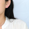 A close-up of a person's ear wearing a small, round, light pink stud earring, with dark hair and a white pleated top against a light blue background.
