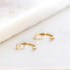 A pair of gold huggie hoop earrings with teardrop-shaped clear gemstones, displayed on a textured white surface.