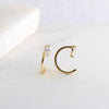 A pair of gold-toned letter C ring earrings with clear heart-shaped gemstones, displayed on a light marble surface.